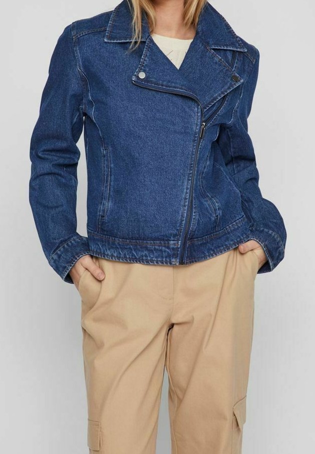 Femme portant une veste biker en jean bleu avec fermeture éclair à l'avant et un pantalon cargo beige, mains dans les poches, debout devant un fond uni.
