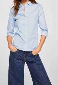 Camicia azzurra chiara con colletto e maniche arrotolate, con un motivo discreto, abbinata a pantaloni a gamba larga in denim scuro a vita alta.