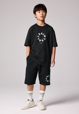 Junge steht mit Händen in den Taschen, trägt schwarzes AllSaints T-Shirt und Shorts, weiße AllSaints Socken und weiße Sneakers vor hellem Hintergrund.