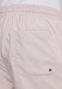 Vaaleanpunaiset shortsit, jotka on valmistettu sileästä kankaasta, jossa on joustava vyötärö ja yksi takatasku, johon on lisätty tummansininen metallirengas.