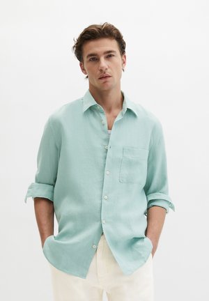 Giovane uomo con le maniche rimboccate, indossa una camicia azzurra a bottoni e pantaloni bianchi, in piedi contro uno sfondo bianco uniforme.