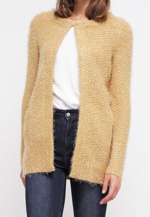 Kvinde iført en beige åben-frontal, blød cardigan over en hvid top kombineret med mørkeblå jeans, stående mod en hvid baggrund.