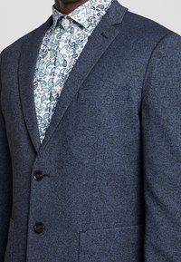 Námořnický modrý upravený blejzr z texturované tkaniny se dvěma černými knoflíky a náprsní kapsou, vrstvený přes vzorovanou bílou košili.