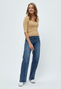 Haut en tricot côtelé beige avec un col en V et des manches trois-quarts, associé à un jean flare en denim bleu taille haute et des baskets blanches.