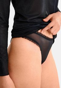 String noir transparent avec des volants en dentelle sur les bords, associé à un haut ajusté à manches longues noir en tissu pailleté.