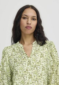 Blusa floreale verde con scollo a V, caratterizzata da maniche a sbuffo e un motivo testurizzato. Sfondo verde chiaro con disegni floreali intricati.