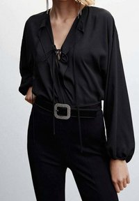 Blouse noire à col en V, avec un détail à cordon de serrage et de longues manches bouffantes. Associée à un pantalon noir ajusté et une ceinture à boucle ornée de bijoux.