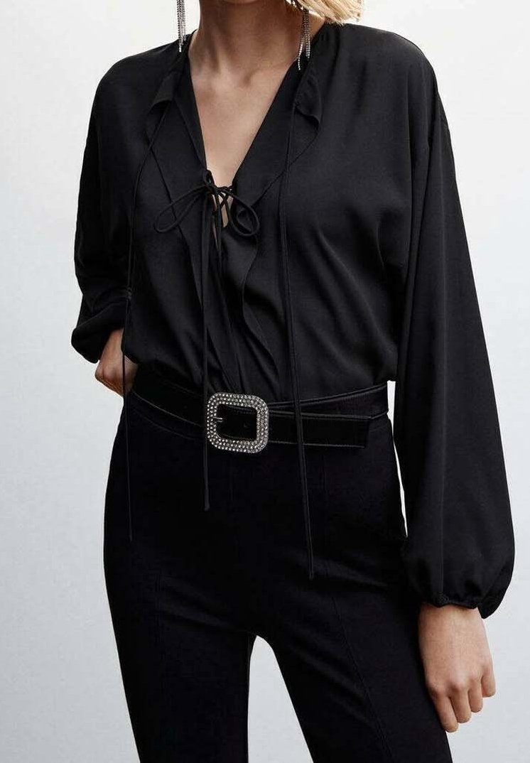 Blouse noire à col en V, avec un détail à cordon de serrage et de longues manches bouffantes. Associée à un pantalon noir ajusté et une ceinture à boucle ornée de bijoux.