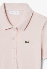 Vaaleanpunainen Lacosten polo-paita, jossa on ribbitekstuuri, klassinen kaulus, kolme nappia ja vihreä krokotiililogo.