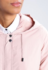 Ljusrosa hooded jacka med tryckknappar, dragsko i halsen och jämn tygtextur, lager över en vit skjorta med krage.