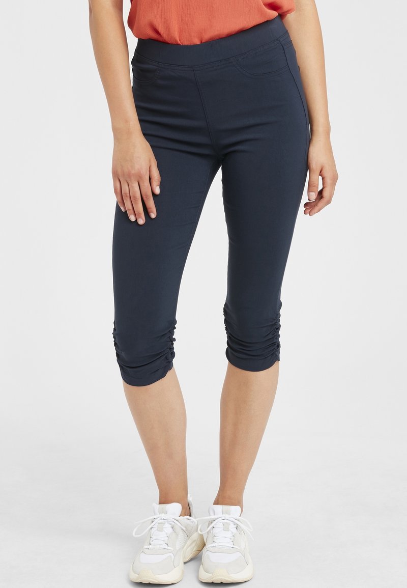 Femme portant des leggings capri bleu marine froncés, des baskets blanches et un haut sans manches orange, debout sur fond blanc.