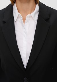 Czarna marynarka z wyłogami i zapięciem na guzik, noszona na białej koszuli z kołnierzykiem. Tkanina wydaje się gładka, o usztywnionej konstrukcji.