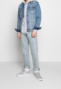Veste en denim avec un lavage bleu clair, sweat-shirt gris en dessous, associée à un pantalon en denim bleu clair retroussé et des baskets grises. Design simple, sans motifs.