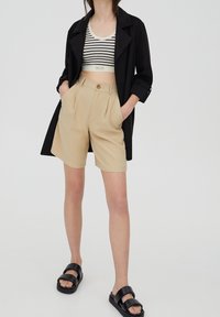 Un abrigo trench negro sobre un top corto a rayas, combinado con pantalones cortos beige de corte ajustado y sandalias negras tipo slide. Se destacan las llanuras y las texturas.