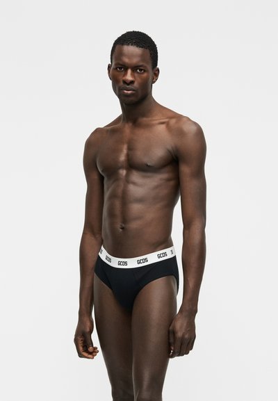 Modèle masculin aux cheveux courts portant un slip noir avec une ceinture blanche marquée "GCDS", posé devant un fond clair uni.