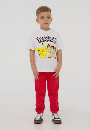 T-shirt bianca in cotone con grafica di Pokémon che presenta Pikachu e Meowth; abbinata a pantaloni jogger rossi e sneakers con accenti scuri.