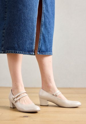 Ben i en blå denimnederdel og beige læder Mary Jane-sko med dobbelte remme, stående på et trægulv.