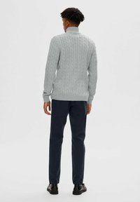 Helles graues Strickpullover mit hohem Kragen, der über ein Zopfmuster und gerippte Ärmelbündchen verfügt, kombiniert mit dunkelblauen Hosen und schwarzen Schuhen.