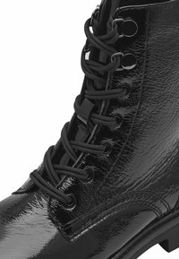 Bottine en cuir verni noir avec une surface lisse et brillante. Présente des lacets ronds noirs et des œillets, avec un design de semelle robuste.