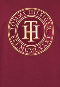 Bordeauxrode stof met een ronde, geborduurde logo met gouden accenten. Het logo bevat "TOMMY HILFIGER" en "EST. MCMLXXV."
