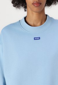 Jasnoniebieska bluza z ribbowanym dekoltem i małą niebieską metką "HUGO". Wykonana z miękkiego materiału, charakteryzuje się luźnym krojem i minimalistycznym designem.