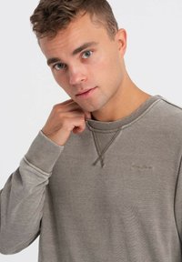 Šedá mikina s texturovaným povrchem, s kulatým výstřihem, žebrovanými manžetami a jemným logem vyšitým na hrudi.