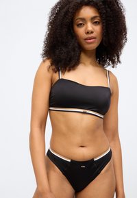 Melns bikini komplekts ar bandeau stila augšdaļu un zemu vidukļa līniju apakšdaļām. Akcentēts ar plānām krēmkrāsas un zelta svītrām gar malām. Gluda tekstūra.