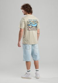 T-paita vaaleasta puuvillasta, jossa on monivärinen Keith Haring -grafiikka selässä, yhdistettynä vaaleansinsisiin farkkushortseihin ja valkoisiin kenkiin, joissa on vihreitä yksityiskohtia.