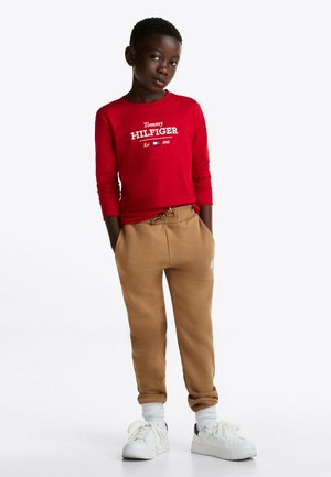 Camicia a maniche lunghe rossa con logo bianco "Tommy Hilfiger", abbinata a pantaloni felpa beige e sneakers bianche con dettagli grigi. Tessuto in cotone testurizzato.