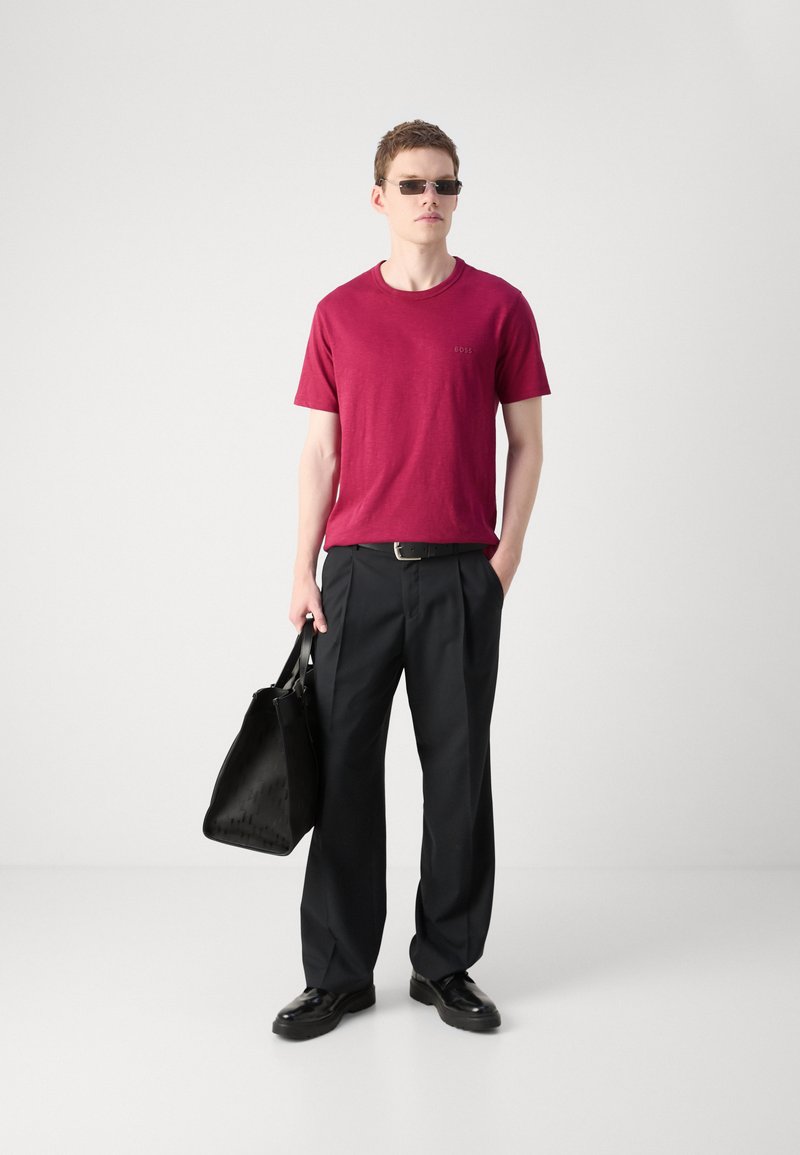 T-shirt rouge à manches courtes avec un logo, pantalon large noir, ceinture noire, lunettes de soleil et sac tote noir, porté avec des chaussures noires.