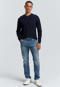 Tengerészkék hosszú ujjú henley ing elülső zsebbel, világoskék farmerrel és fehér tornacipővel párosítva. Hétköznapi viseletre alkalmas laza öltözet.