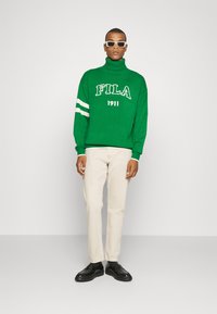 Grün gestrickter Rollkragenpullover mit weißem "FILA 1911"-Text und drei weißen Streifen an den Ärmeln. Kombiniert mit cremefarbenen Hosen und schwarzen Schuhen.