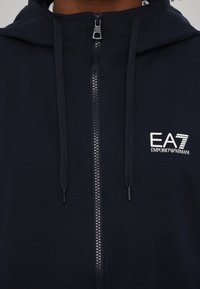 Jūras spēku zip-aizdare pelēks džemperis, izgatavots no kokvilnas maisījuma, ar mazu baltu EA7 logo uz krūšu un kapuci ar aukliņu. Gluda tekstūra un regulāra piegriezuma.