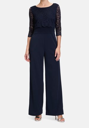 Femme portant une combinaison bleu marine avec des manches trois-quarts en dentelle et un pantalon à jambes larges, assortie à des sandales argentées à talons hauts.
