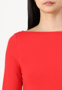 Crveni, uski gornji dio s brodskim izrezom i dugim rukavima. Glatka, rastezljiva tkanina bez vidljivih uzoraka ili akcenta.