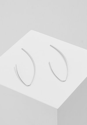 Paire de boucles d'oreilles fines en fil d'argent courbé, présentée sur un cube blanc sur un fond gris neutre.
