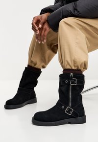 Crne gležnjače od brušene kože s ravnim gumenim potplatom, bočnim patentnim zatvaračem i podesivim remenom s kopčom. Meka tekstura i casual dizajn.