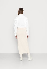 Frau mit langen braunen Haaren steht vor einer grauen Wand und trägt ein weißes Langarmshirt, einen beigen langen Rock und weiße Stiefel.