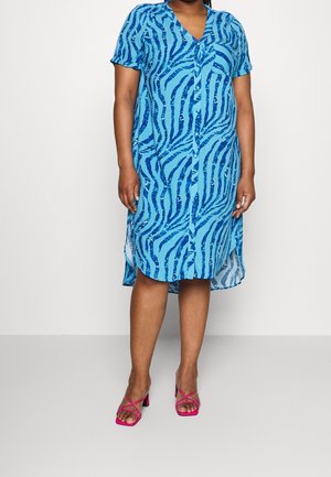 Femme portant une robe boutonnée bleu à rayures zébrées à manches courtes avec des sandales à talons roses à brides, debout sur un sol gris.