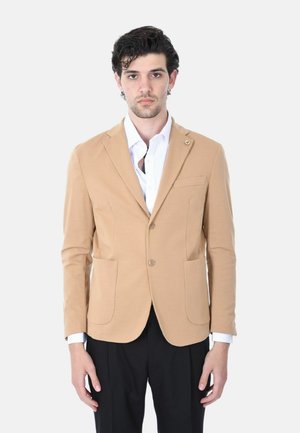 Uomo con blazer beige sopra una camicia bianca e pantaloni neri, in piedi davanti a uno sfondo chiaro uniforme, rivolto verso la fotocamera.