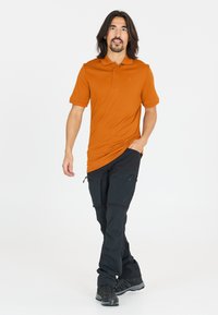 Orange polo med korta ärmar, kombinerat med svarta cargobyxor med fickor som har dragkedjor. Modell som bär mörka skor, står framför en vit bakgrund.