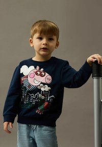 Niño pequeño con un sudadera azul marino con un cerdo y un dinosaurio de dibujos animados, sosteniendo un poste plateado y negro frente a un fondo liso.