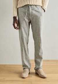 Pantalons gris clair texturés avec une coupe décontractée et une taille à cordon. Portés avec des chaussures à enfiler beiges, debout sur un sol en bois.