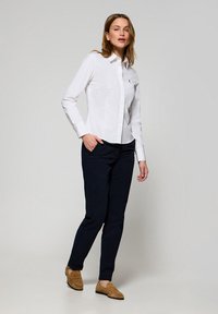 Camicia bianca con bottoni, maniche lunghe, abbinata a pantaloni blu scuro e scarpe marrone chiaro. La camicia presenta una vestibilità slim e un colletto classico.