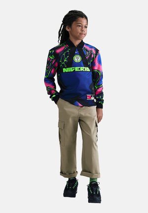 Dreng med langt hår iført farverig Nigeria fodboldtrøje, beige cargobukser, grønne sokker og sorte sneakers, står med hænderne i lommerne.