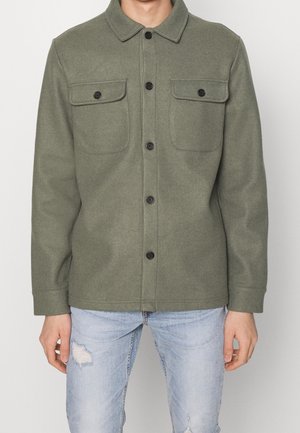 Man met een olijfgroene jas met knopen, twee borstzakken en lichtblauwe gerafelde spijkerbroek, tegen een effen achtergrond.