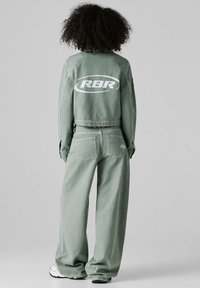 Persona con cabello rizado viste una chaqueta verde con la marca RBR y pantalones sueltos a juego, de pie de espaldas sobre un fondo liso.