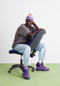 Fialové tenisky, šedé tričko s krátkým rukávem, modré džíny a fialový kulich; sedí na modré kancelářské židli na zelené podlaze.