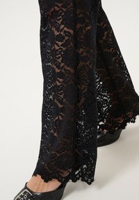 Zwarte kanten broek met bloemenpatroon, doorschijnende textuur en een schulpzoom. Gecombineerd met zwarte lakleren schoeisel.