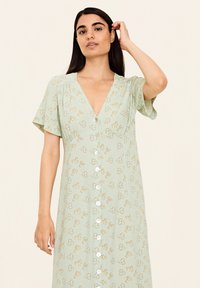 Mintgrünes Kleid mit V-Ausschnitt, kurzen, fließenden Ärmeln und durchgehender Knopfleiste. Es hat ein florales Muster mit gelben Akzenten und einer strukturierten Stoffqualität.